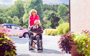 a nurse next door caregiver pushes an elderly client in a wheelchair towards his house