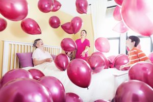 older person in bed surrounded by nurse, grandson and lots of pink balloons to focus on happiness