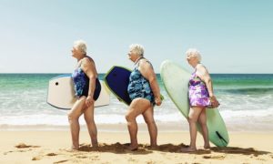 3 older women on the beach with their surfboards under their arms