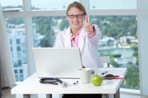 Young female wearing lap coat using laptop showing thumbs up sign sitting behind desk
